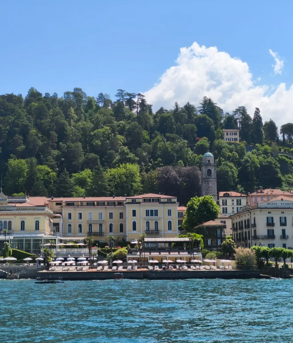 Lakefront view of elegant historic buildings and grand hotels along the shore, with a wooded hillside, blue sky, and fluffy white clouds in the background.